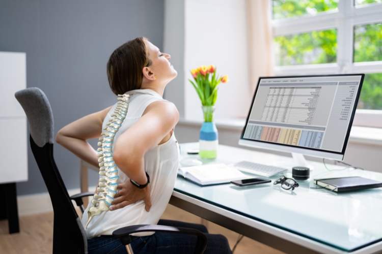 Žena sedi za radnim stolom i drži donji deo leđa zbog bola, dok ilustracija kičme prikazuje posledice koje može da izazove loše sedenje tokom rada za računarom.