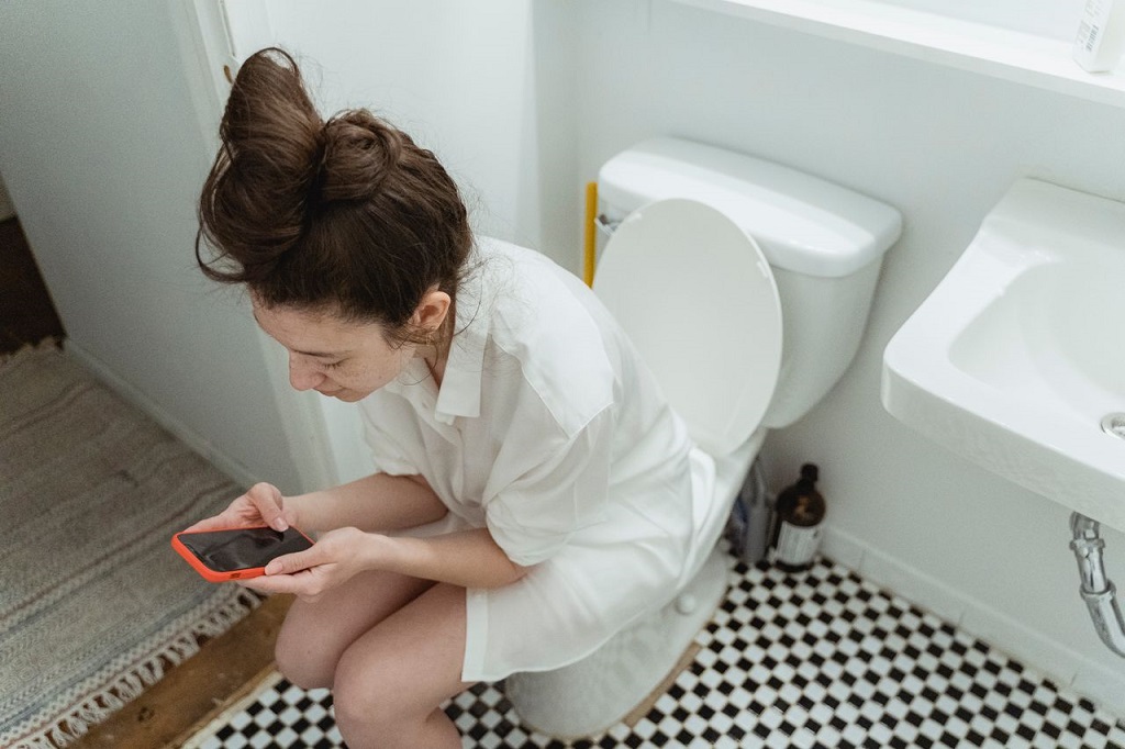 Devojka u toaletu sedi na wc šolji sa telefonom u ruci